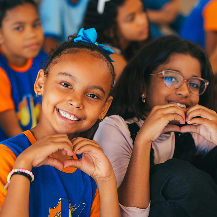 Duas meninas sorrindo em primeiro plano. Uma à frente, a outra com óculos, fazendo um coração com as mãos.