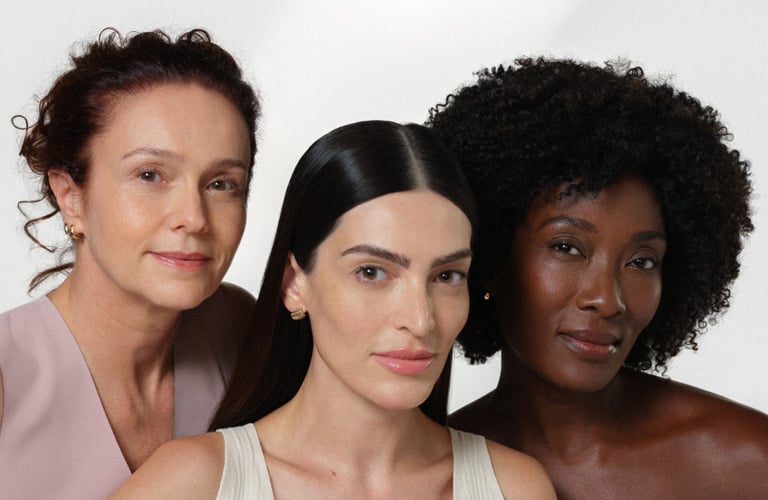Foto com fundo branco e três mulheres posando lado a lado, com os rostos parcialmente cobertos, vestindo roupas em tons claros.