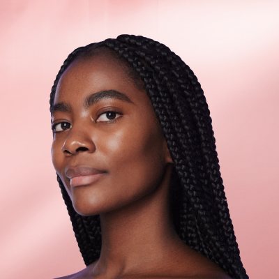 Mulher negra jovem, com tranças box braids longas e pele brilhante com aspecto natural e saudável. Ela sorri levemente e olha para a câmera.