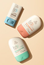 Três Protetores Solares Faciais FPS 50 Natura dispostos em diagonal. Todos têm embalagem branca, mas a base do stick é azul e as tampas dos demais são verde e laranja. Fundo em tom de creme.