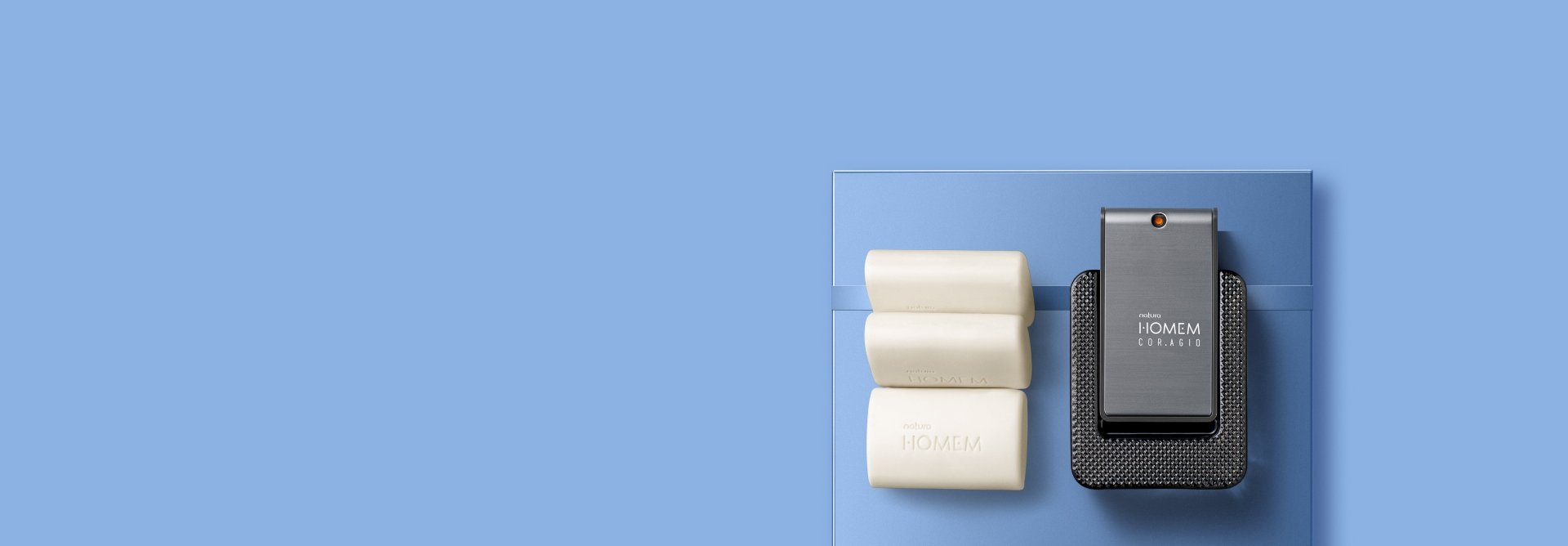 Sabonetes em barra brancos Natura Homem e frasco de fragrância Homem Cor.agio, sobre caixa azul com faixa, em fundo azul.