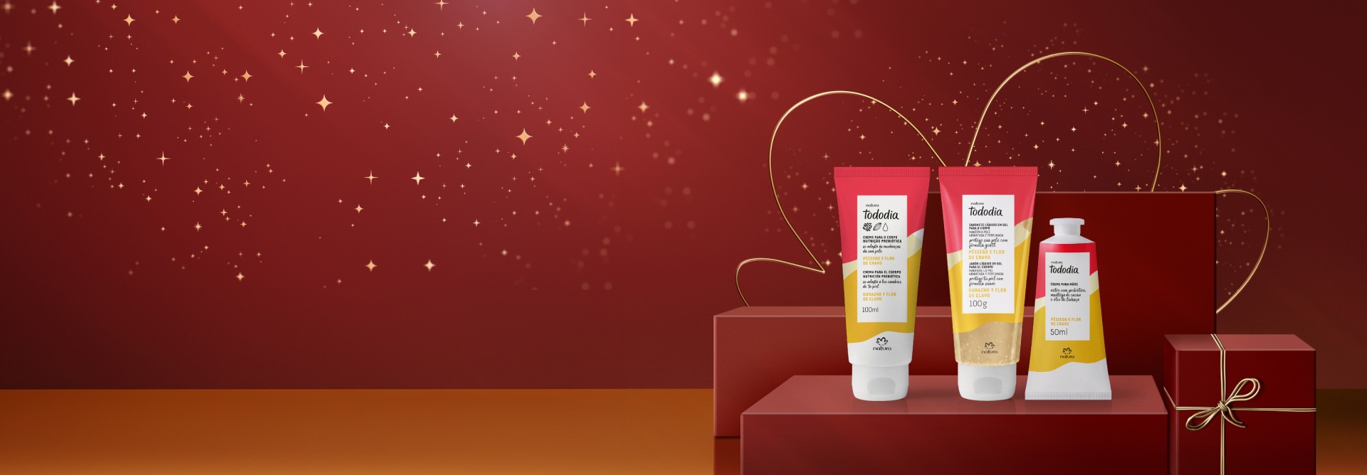 Presente Natura Tododia Pêssego e Flor de Cravo, com Creme Hidratante para o Corpo, Creme para as Mãos e Sabonete Líquido em Gel, sobre caixas em fundo vermelho. Embalagens bisnaga em branco, amarelo e rosa.