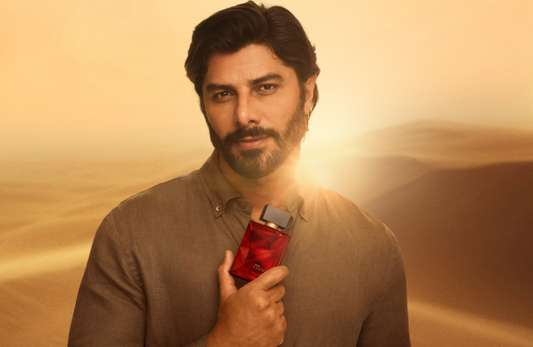 Foto em cenário de dunas ao pôr do sol. Homem segura o frasco vermelho do perfume Essencial próximo ao corpo.
