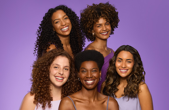 Foto de cinco mulheres sorrindo com diferentes tipos de cabelos cacheados e crespos, dispostas em duas fileiras. Fundo roxo.