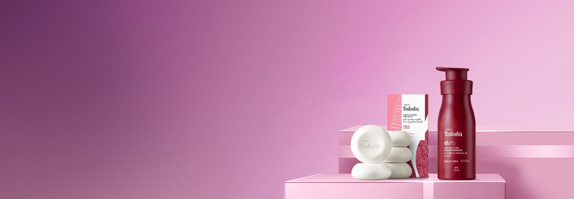 Creme para o Corpo e Sabonete em Barra Puro Vegetal da linha Tododia Cereja e Avelã sobre caixas rosas com laços na mesma cor. Fundo liso rosa.