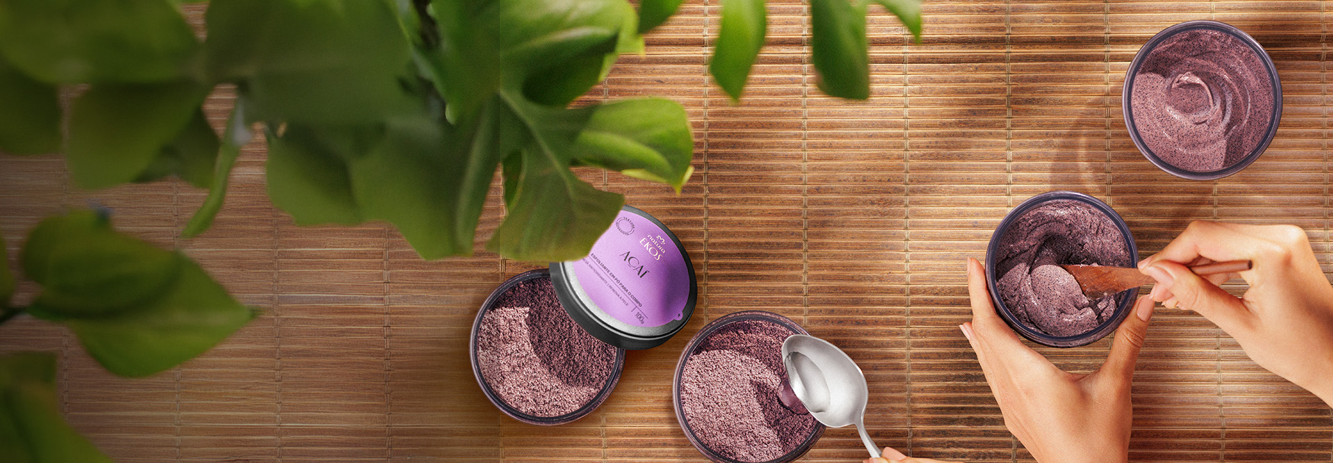 Sobre bancada em madeira, potes abertos do Esfoliante em Pó para o Corpo Ekos Açaí. Acima do pote esquerdo há uma folha, ao centro uma colher despejando textura transparente e à direita mãos brancas misturam produto com colher de madeira.