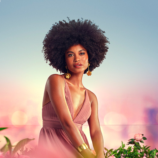 Modelo negra de cabelos crespos usa vestido longo rosê e brincos dourados. Ela posa para a câmera em meio a flores e folhagens em fundo rosado.
