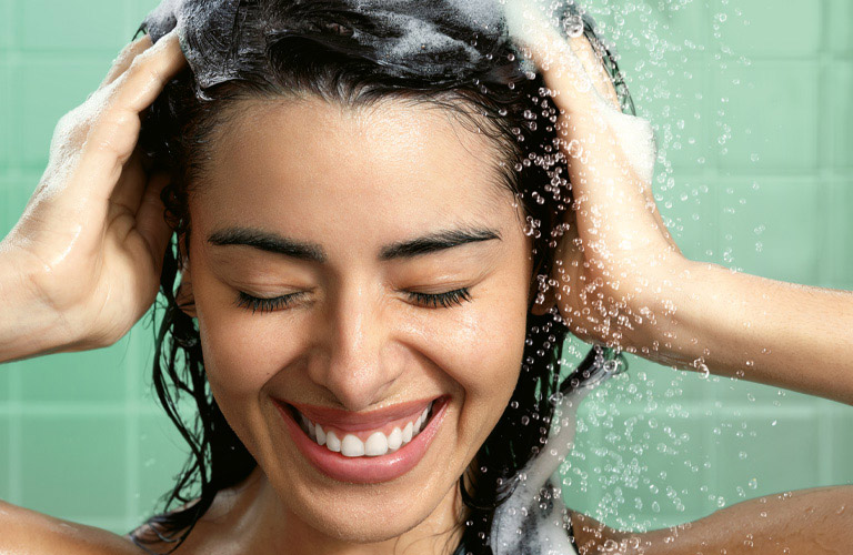 Foto de uma mulher no banho, com as mãos no cabelo e água caindo. Ao fundo, azulejos verdes.