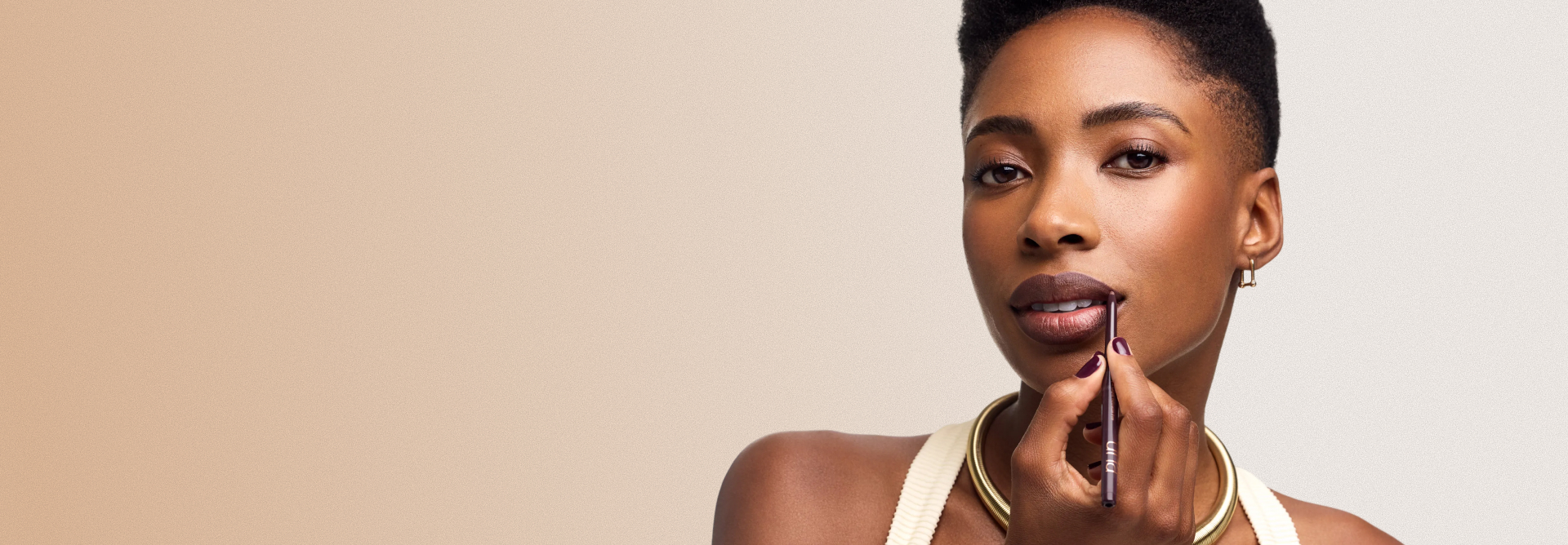 Mulher de pele negra e cabelos curtos aplica o Lápis Labial Retrátil PRO Una nos lábios. Ela veste uma blusa clara e um colar dourado, sobre um fundo bege gradiente.