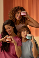 Três mulheres de diferentes etnias e tipos de cabelo (cacheado, liso e com franja, e ondulado) sorrindo, segurando potes de Máscara de Tratamento Natura Tododia em cores diferentes (rosa, laranja e azul) para o cronograma capilar.