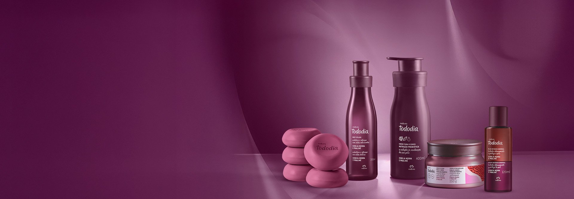 Linha Tododia Cereja Negra e Praliné com 5 sabonetes redondos, 1 frasco de body splash, 1 pump de creme, 1 pote de creme veludo e 1 frasco de óleo corporal. Embalagens roxas em fundo roxo iluminado.
