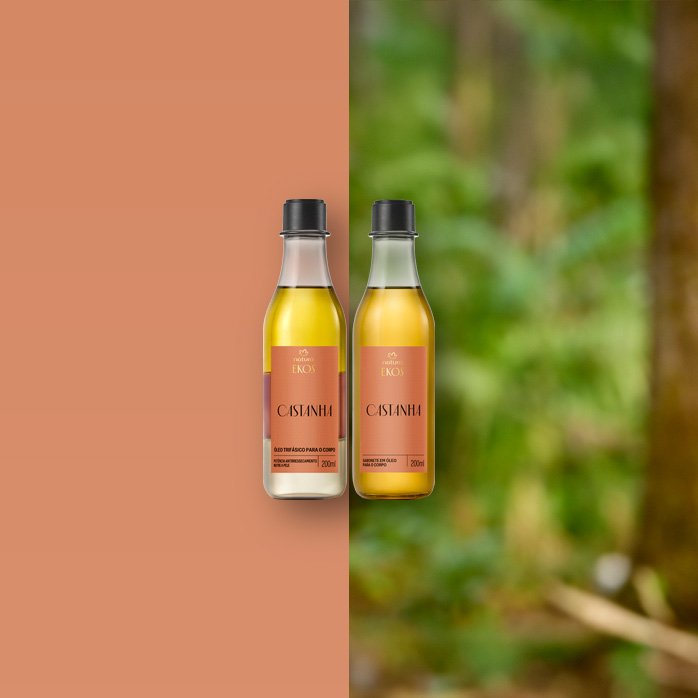 Dois frascos de vidro do Óleo Trifásico Ekos Castanha, com tampa preta e rótulo marrom, estão sobre um galho grosso em meio a folhas verdes e troncos de árvore na floresta.