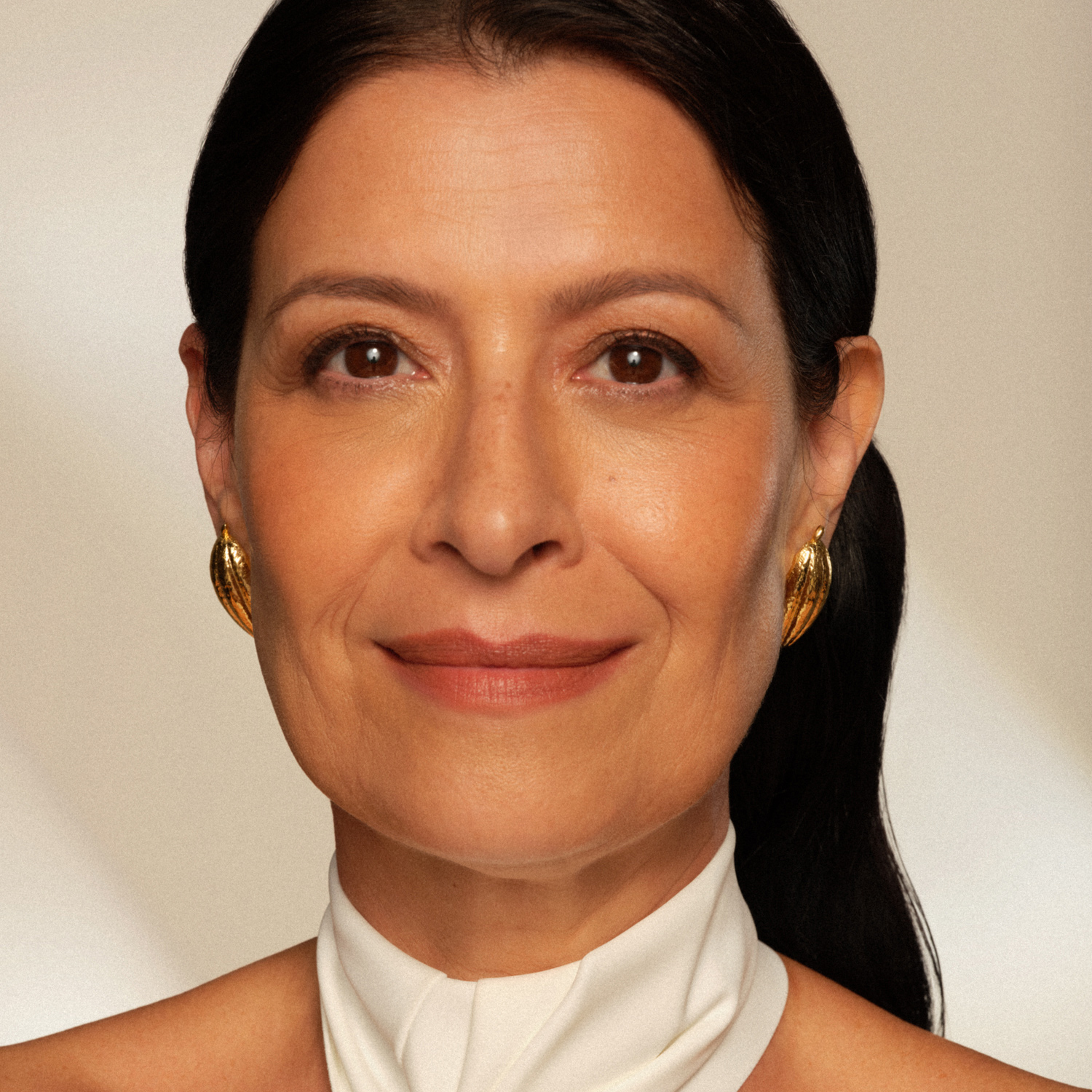 Mulher de pele madura branca com olhos castanhos e cabelo castanho preso olha fixamente para a câmera com expressão de confiança e tranquilidade. Ela veste branco e usa brincos dourados discretos. 