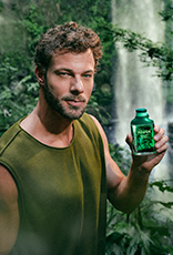 Modelo de cabelos e barba castanho-claros usa regata verde-musgo e segura o novo Kaiak Aventura Intensa Masculino nas mãos. Ao fundo, destaca-se a imagem de uma cachoeira em meio à floresta.
