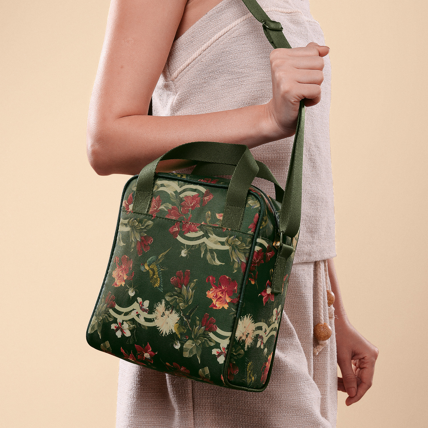 Mulher segurando bolsa de ombro verde com estampa de flores e pássaros. Corte da imagem abaixo do ombro com fundo bege.