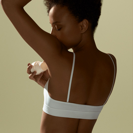 Mulher de costas aplicando desodorante roll-on Natura, em fundo verde-claro.