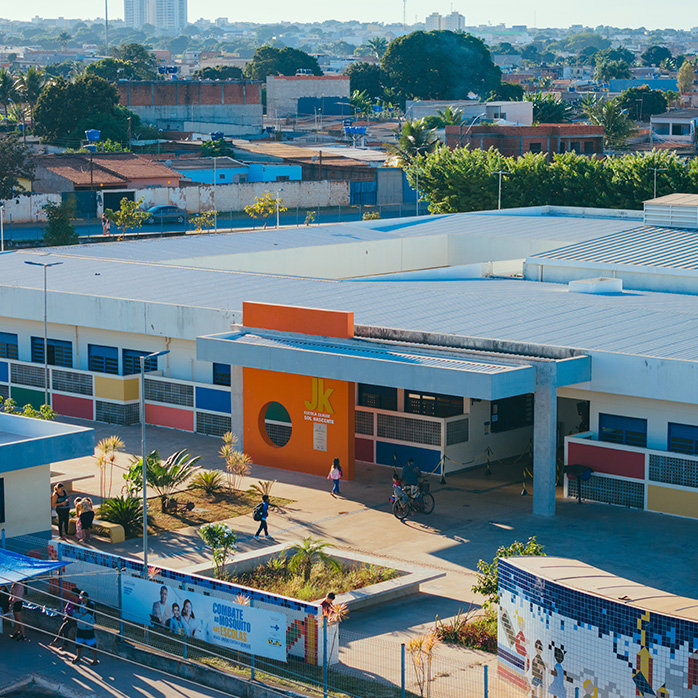 Vista aérea da escola JK com fachada colorida, árvores ao redor e pessoas circulando na entrada principal.