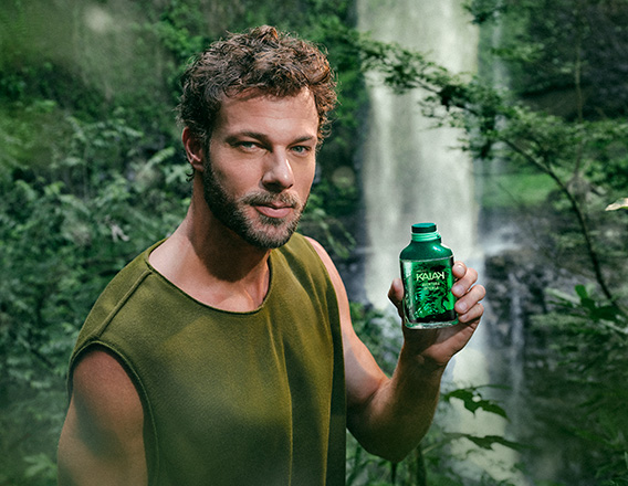 Modelo de cabelos e barba castanho-claros usa regata verde-musgo e segura o novo Kaiak Aventura Intensa Masculino nas mãos. Ao fundo, destaca-se a imagem de uma cachoeira em meio à floresta.