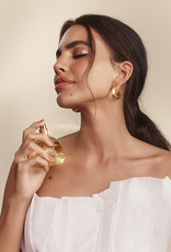 Foto de mulher branca de cabelos castanhos presos em um rabo-de-cavalo, aplicando o Perfume Natura Aura Alba no pescoço.