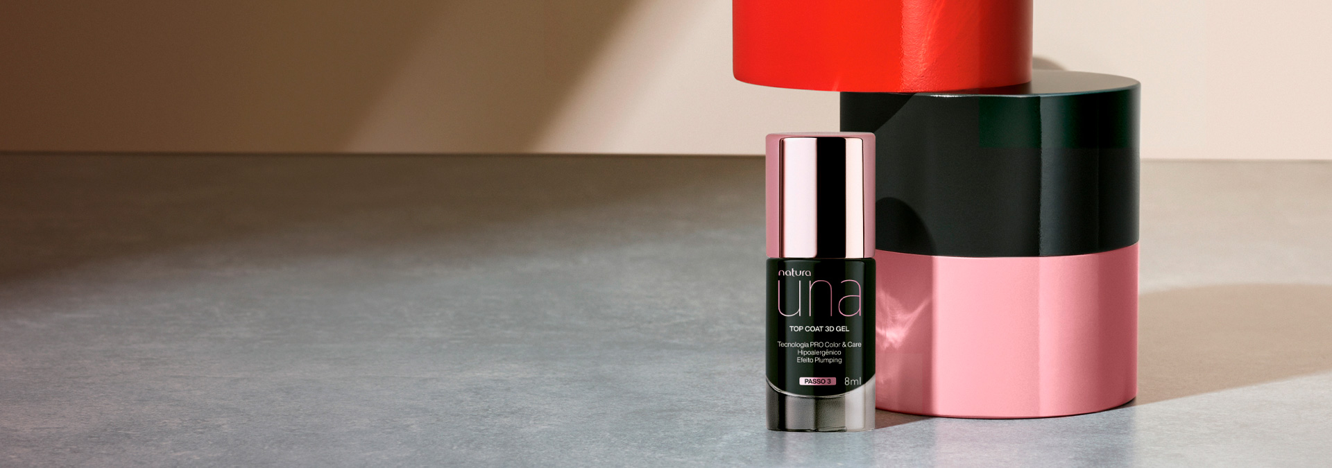 Fundo cinza e ao lado direito temos o top coat para unhas Una com elementos rosa, preto e vermelho ao fundo