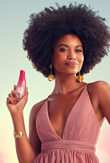Modelo negra de cabelos crespos usa vestido longo rosê com brincos e bracelete dourados. Ela sorri para a câmera e exibe a fragrância Natura Luna Divina na mão direita.