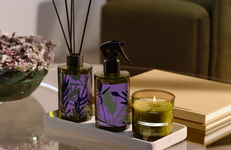 Vela aromática, Spray de ambiente e Difusor da linha Bothânica juntos em cima de uma mesinha de vidro com um sofá marrom atrás