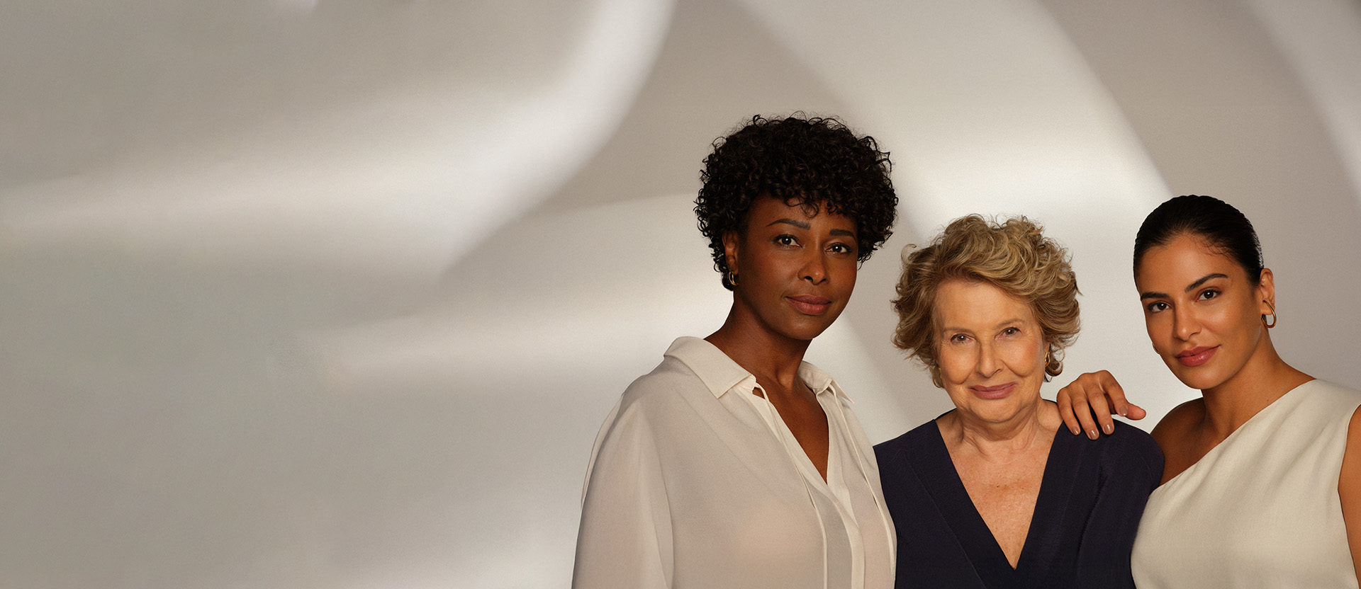 Três mulheres de diferentes gerações e etnias posam juntas sorrindo suavemente sobre um fundo neutro e iluminado.