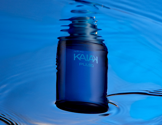 Frasco azul-escuro da fragrância Kaiak Pulso flutuando em água azul, com reflexos ondulados.
