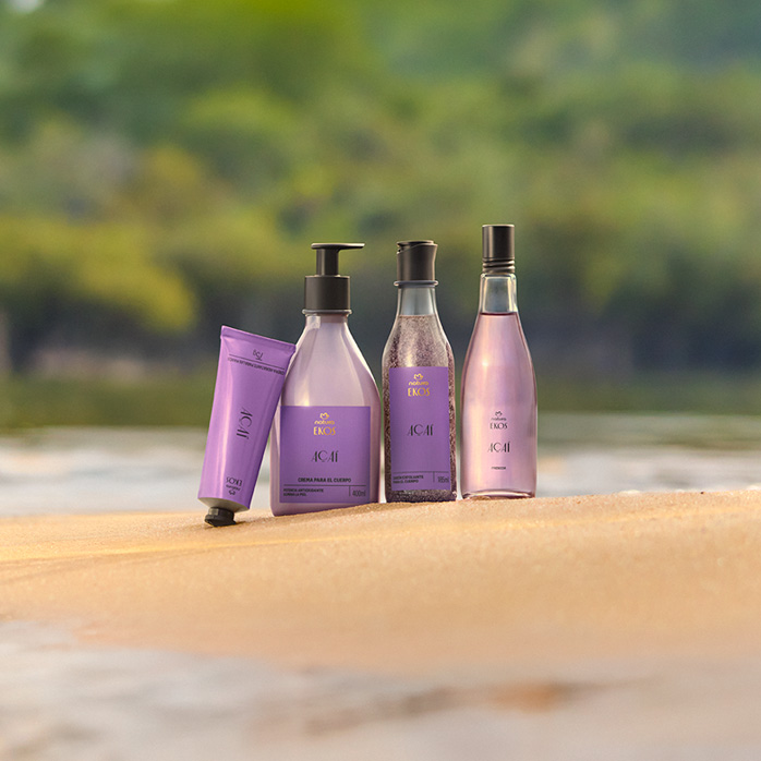 Quatro produtos Natura Ekos Açaí em embalagens lilás estão dispostos sobre a areia, com fundo de rio e vegetação desfocada ao fundo, transmitindo frescor e natureza.
