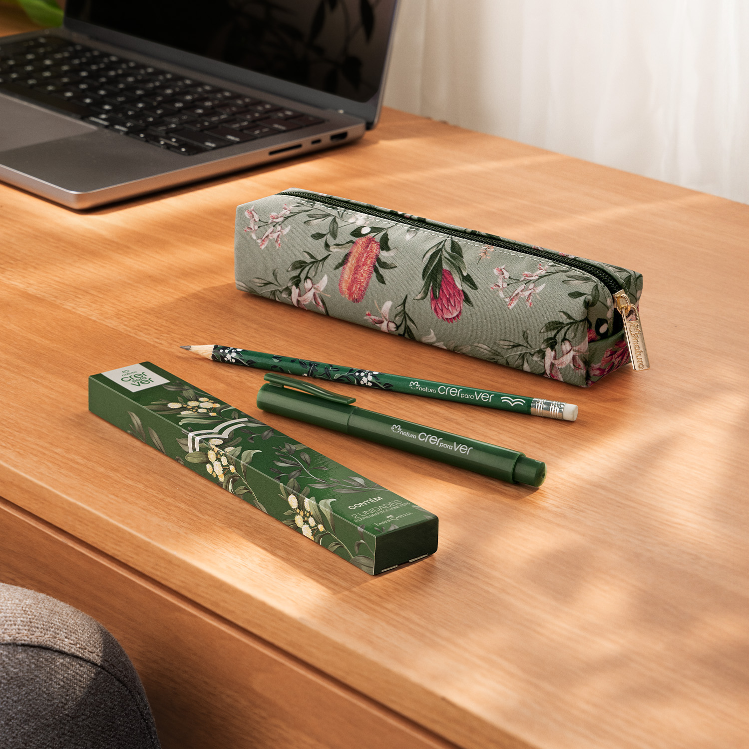 Sobre escrivaninha de tom amadeirado estão uma caneta, um lápis, uma caixa de presente, um estojo florido e um notebook preto. Os itens de papelaria são todos verdes e têm estampa floral combinando. 
