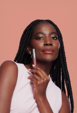 Foto de mulher negra de tranças segurando um batom Natura Una próximo ao rosto, em fundo rosa-claro.