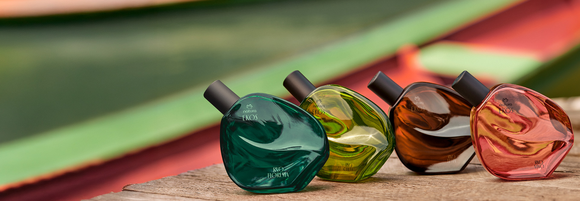 Os quatro frascos de Desodorante Colônia Natura Ryo Floresta, Ryo Chuva, Ryo Festa e Ryo Vivo, aparecem alinhados sobre madeira, com o rio colorido ao fundo levemente desfocado.