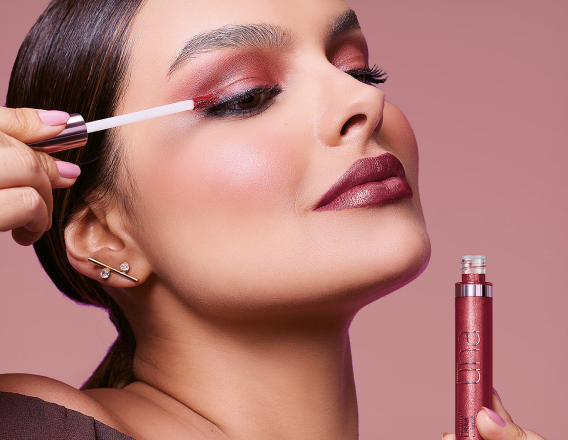 Foto de mulher com cabelos castanhos e presos aplicando o produto Natura Una nas pálpebras, em fundo rosa-claro.