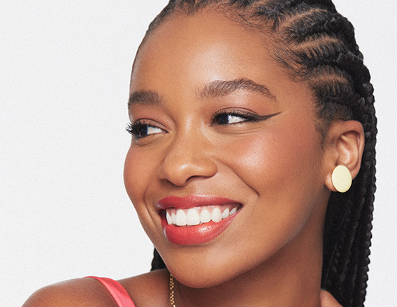 Mulher negra de cabelo escuro trançado sorri olhando para o lado. Ela usa batom vermelho, delineado preto, brinco e colar dourados e uma blusa de alça rosa. Fundo branco.