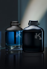 Frascos das fragrâncias K em azul e K Max em preto. Fundo escuro com reflexos e um feixe de luz suave ao fundo.