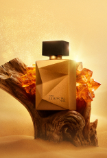 Deo Parfum Essencial Mirra Masculino em frasco facetado dourado com tampa preta. Está em fundo areia sobre um tronco de madeira escura e pedras âmbar ao redor.