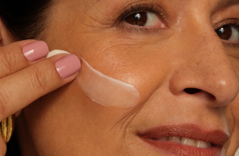 Mulher branca de perfil passando sérum na bochecha direita com dois dedos