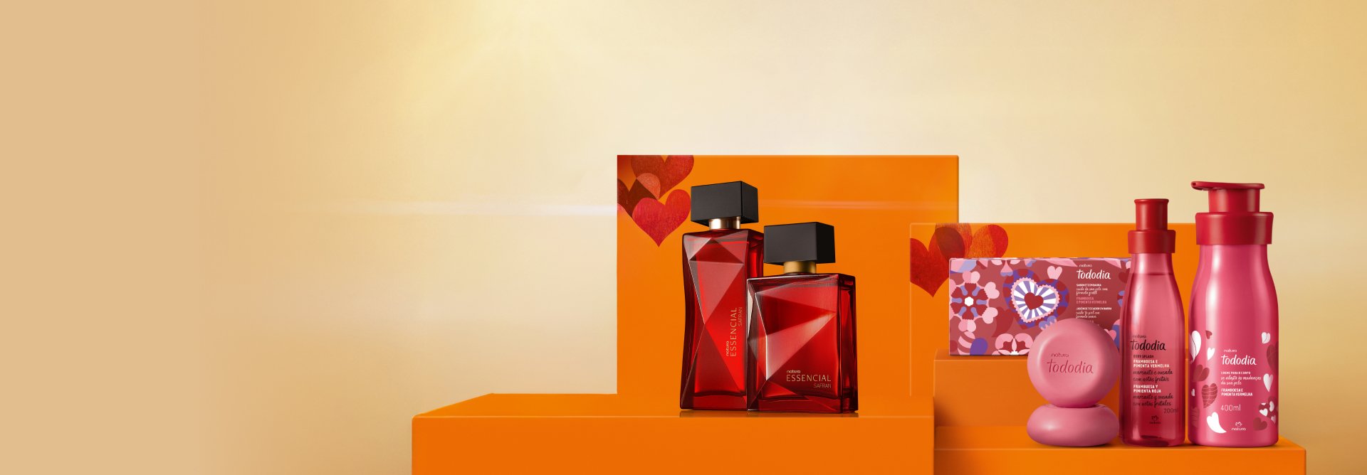 2 frascos vermelhos de fragrâncias Essencial Safran e 3 produtos vermelhos de Tododia Framboesa e Pimenta Vermelha. Estão sobre caixas de presente laranja com corações vermelhos, em fundo amarelo.
