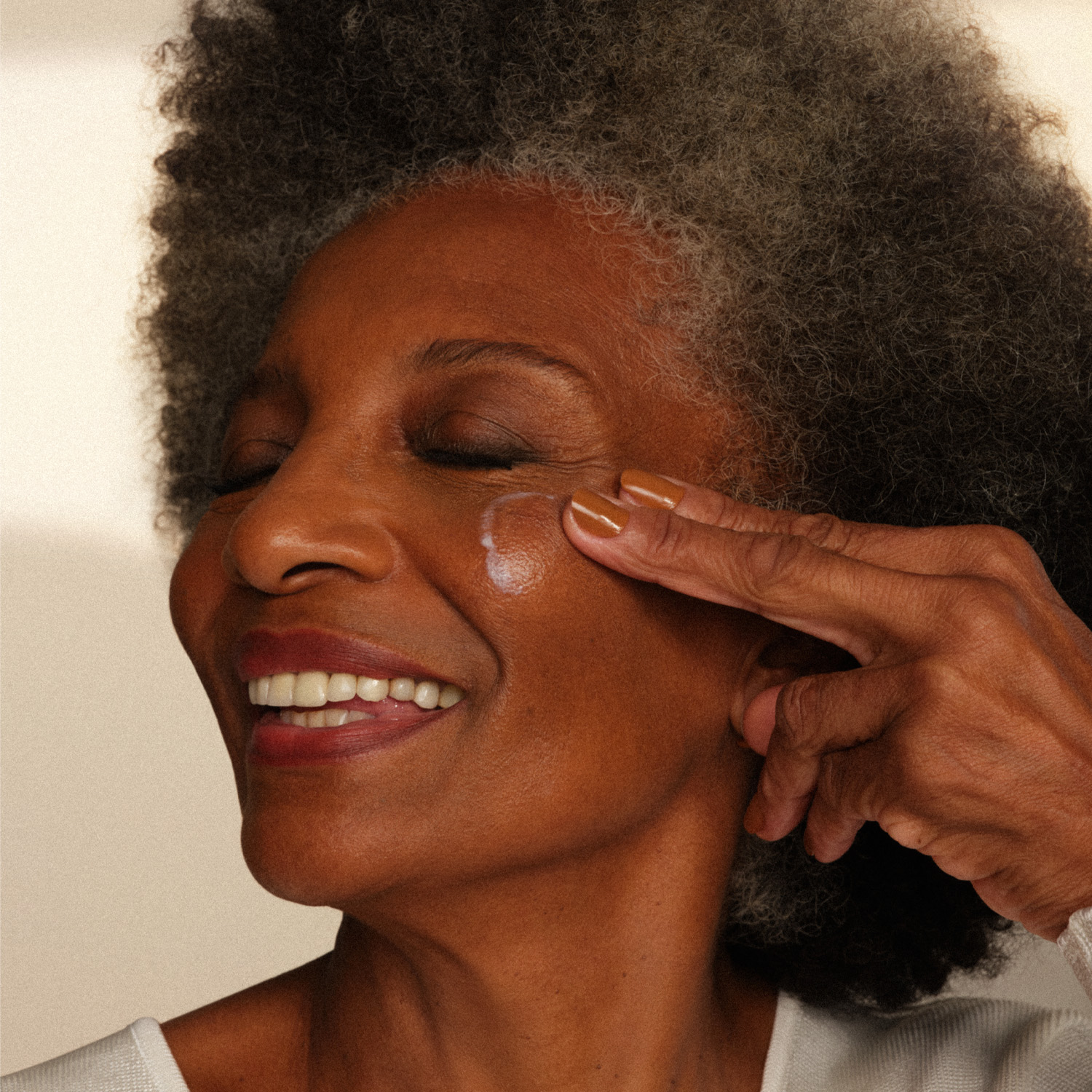Foto de uma modelo de pele madura sorridente. Ela tem cabelos curtos e loiros, usa brincos dourados e blusa preta.
