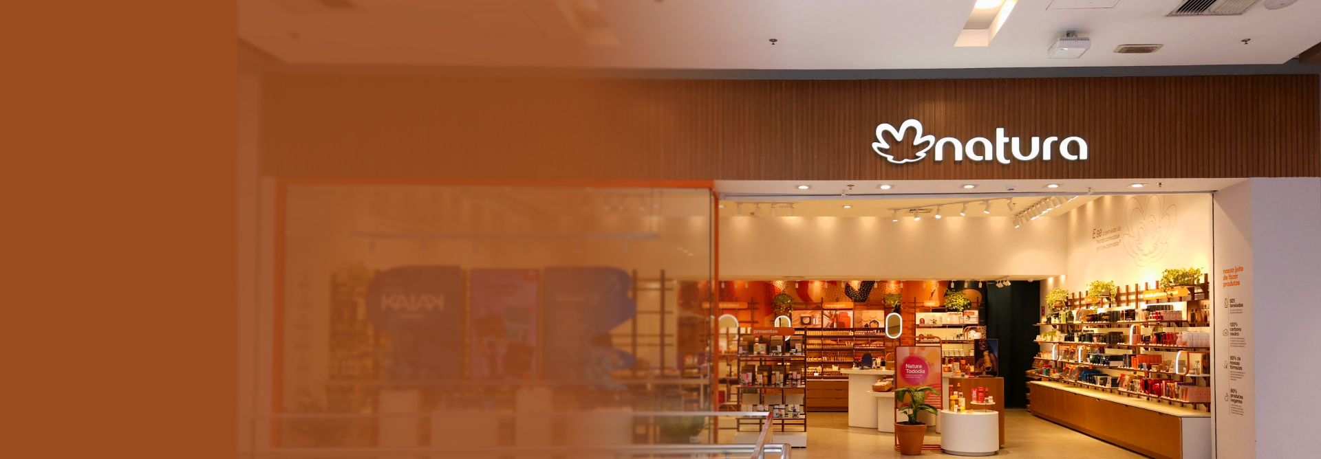 Imagem em degradê laranja revela uma loja física da Natura. Na entrada, logo da Natura iluminado sobre fachada de madeira e vitrine decorada. Dentro da loja, produtos organizados por categorias.