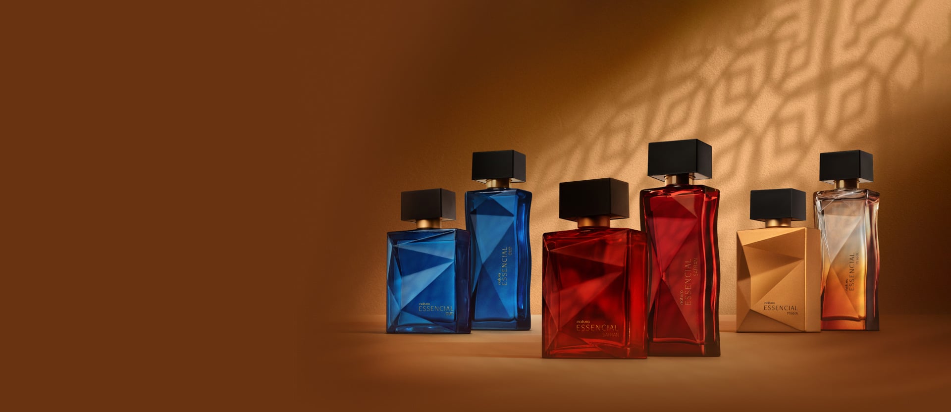 6 frascos de fragrâncias sendo 2 azuis de Essencial Oud, 2 vermelhos de Essencial Safran e 2 alaranjados de Essencial Mirra, todos com tampas pretas. Estão em fundo marrom com sombra geométrica. 