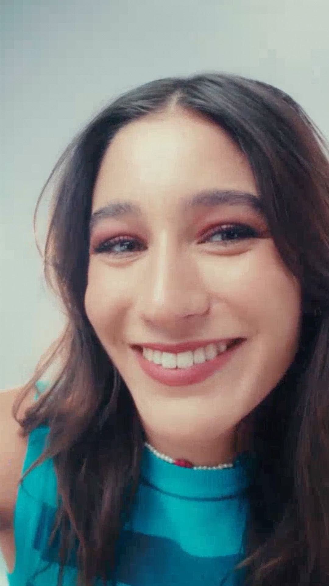 A modelo exibe alguns itens de Natura Faces, incluindo uma sombra, uma máscara de cílios e uma máscara para olhos e sobrancelhas. Diante do espelho, ela aplica os produtos no rosto, criando uma maquiagem vibrante e cheia de cor.