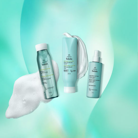 Três produtos Natura Tododia Maçã Verde e Aloe Vera para cabelos, em embalagens verde-água: um shampoo, um condicionador e um spray, com suas respectivas texturas. Dispostos em fundo turquesa com espuma branca.