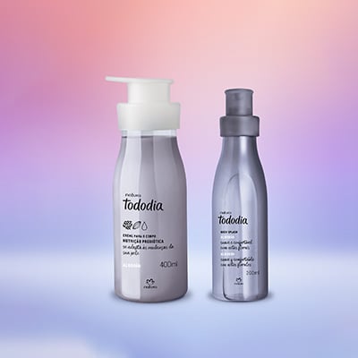 Creme para o Corpo Tododia Algodão em frasco transparente com tampa pump branca e Body Splash Tododia Algodão em frasco lilás com tampa lilás. Produtos sobre fundo degradê rosa e lilás.