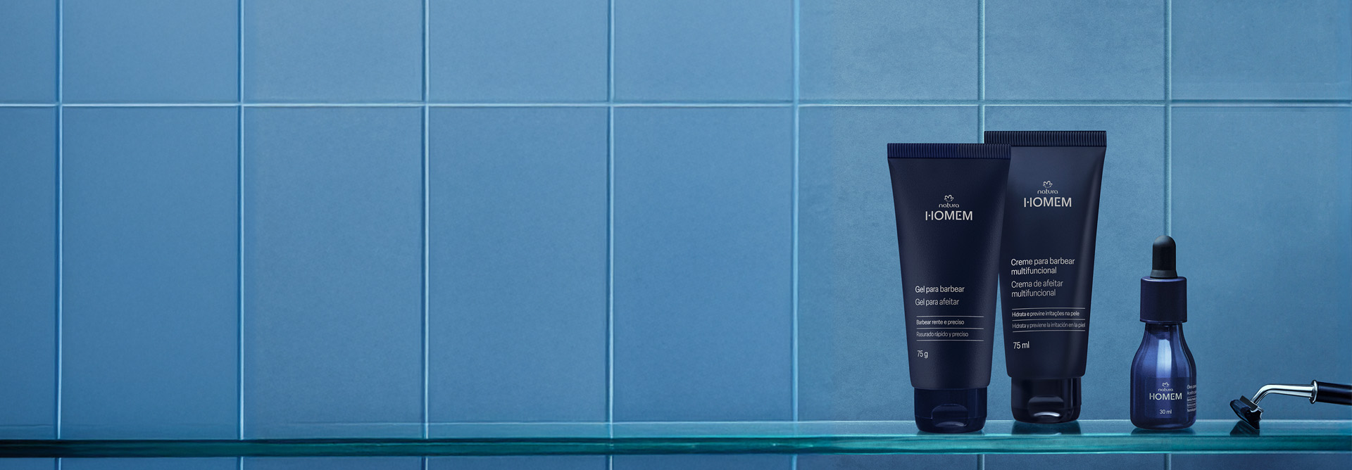 Natura Homem Gel para Barbear, Creme para Barbear Multifuncional e Óleo para Barba, com frascos azul-escuros e tampas pretas, destacam-se sobre prateleira de vidro em fundo azulejado azul-celeste. Ao lado, lâmina de barbear.