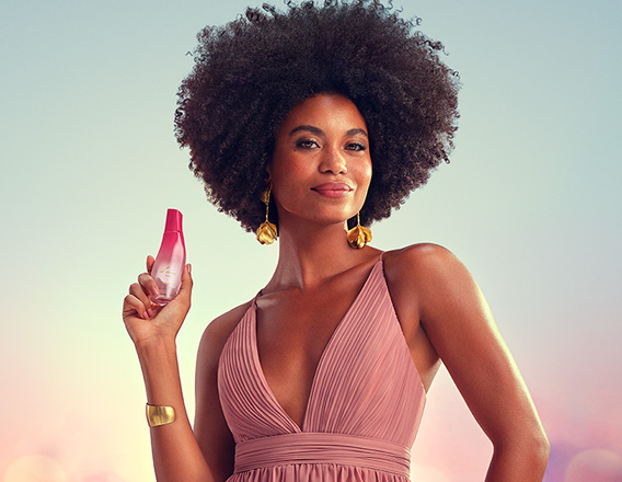 Modelo negra de cabelos crespos usa vestido longo rosê com brincos e bracelete dourados. Ela sorri para a câmera e exibe a fragrância Natura Luna Divina na mão direita.