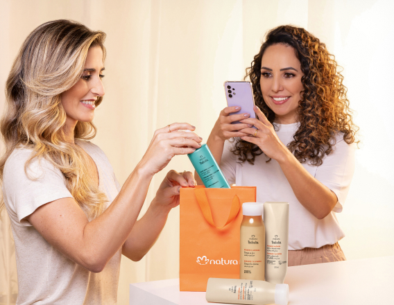 Imagem mostra mulher loira de pele clara colocando itens Natura Tododia para Cabelos em uma sacola laranja da Natura. Uma mulher morena de pele clara e cabelos cacheados filma a cena com o celular.