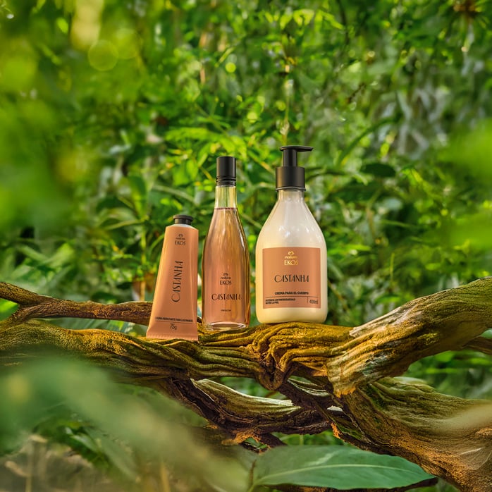 Kit Natura Ekos Castanha com bisnaga bege de polpa hidratante para mãos, frasco transparente de óleo trifásico e pump de creme corporal, apoiados em galho, com fundo de floresta verde.