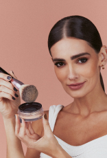 Foto de mulher de cabelos castanhos e presos segurando um produto Natura Una com um pincel, em fundo rosa-claro.