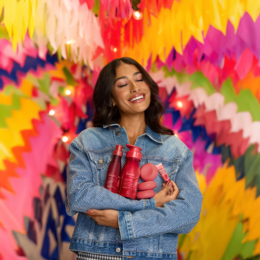 Mulher de cabelos castanhos, usando jaqueta jeans, segura produtos da linha Natura Tododia em fundo com bandeirinhas juninas desfocadas.